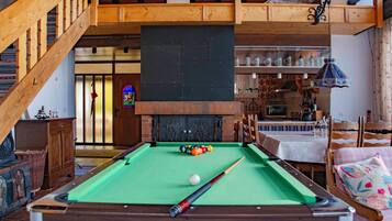 A pool table with balls, a billiard cue, and a fireplace in a room with wooden beams and a staircase.