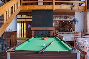 A pool table with balls, a billiard cue, and a fireplace in a room with wooden beams and a staircase.