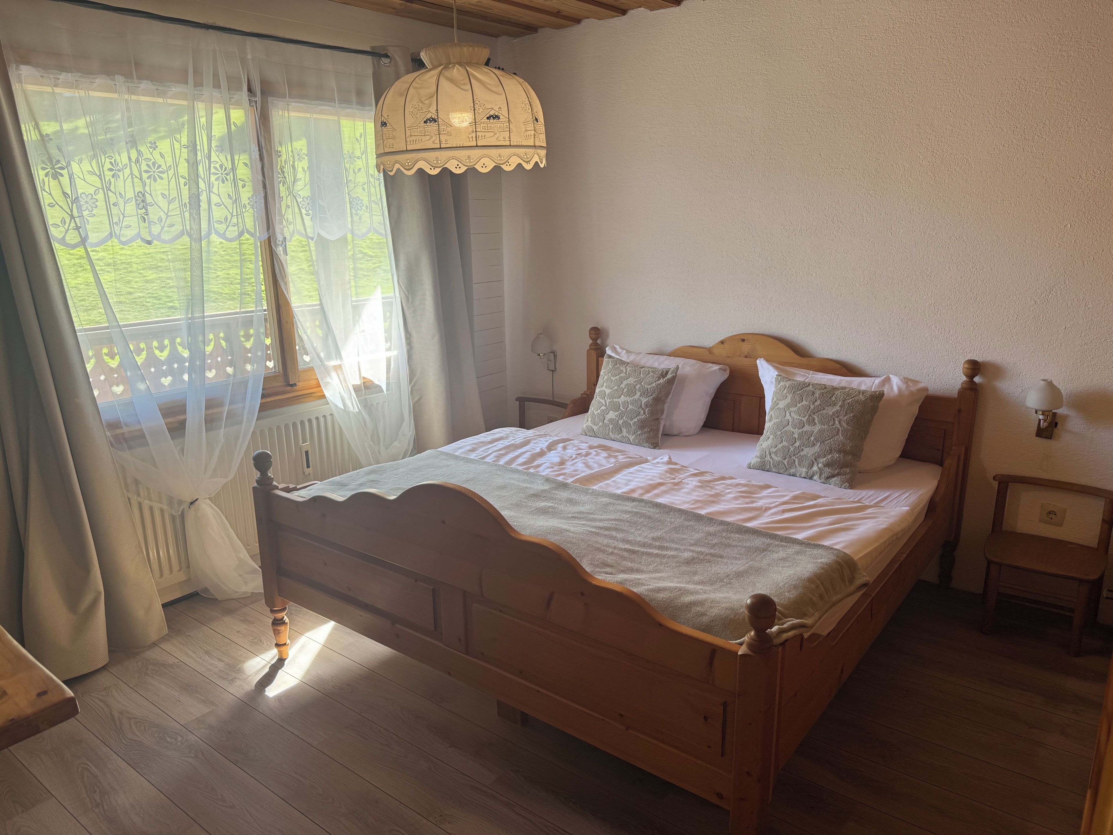 A wooden bed with a white bedspread and pillows, a nightstand with a lamp, and a view of a green landscape through sheer curtains.