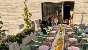 An outdoor dining area with a long wooden table set with plates, cutlery, and glasses.