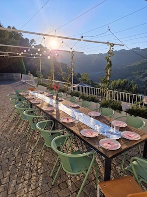 Un área de comedor al aire libre con una larga mesa dispuesta para cuatro personas, rodeada de sillas verdes, con vistas a un paisaje montañoso.