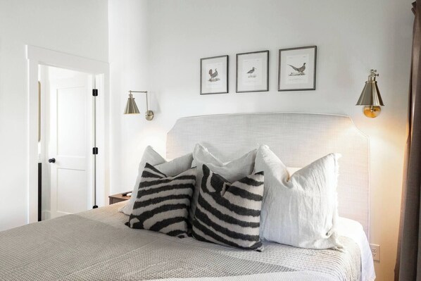 Una cama bien hecha con almohadas a rayas, una lámpara de noche e ilustraciones enmarcadas de pájaros en la pared.