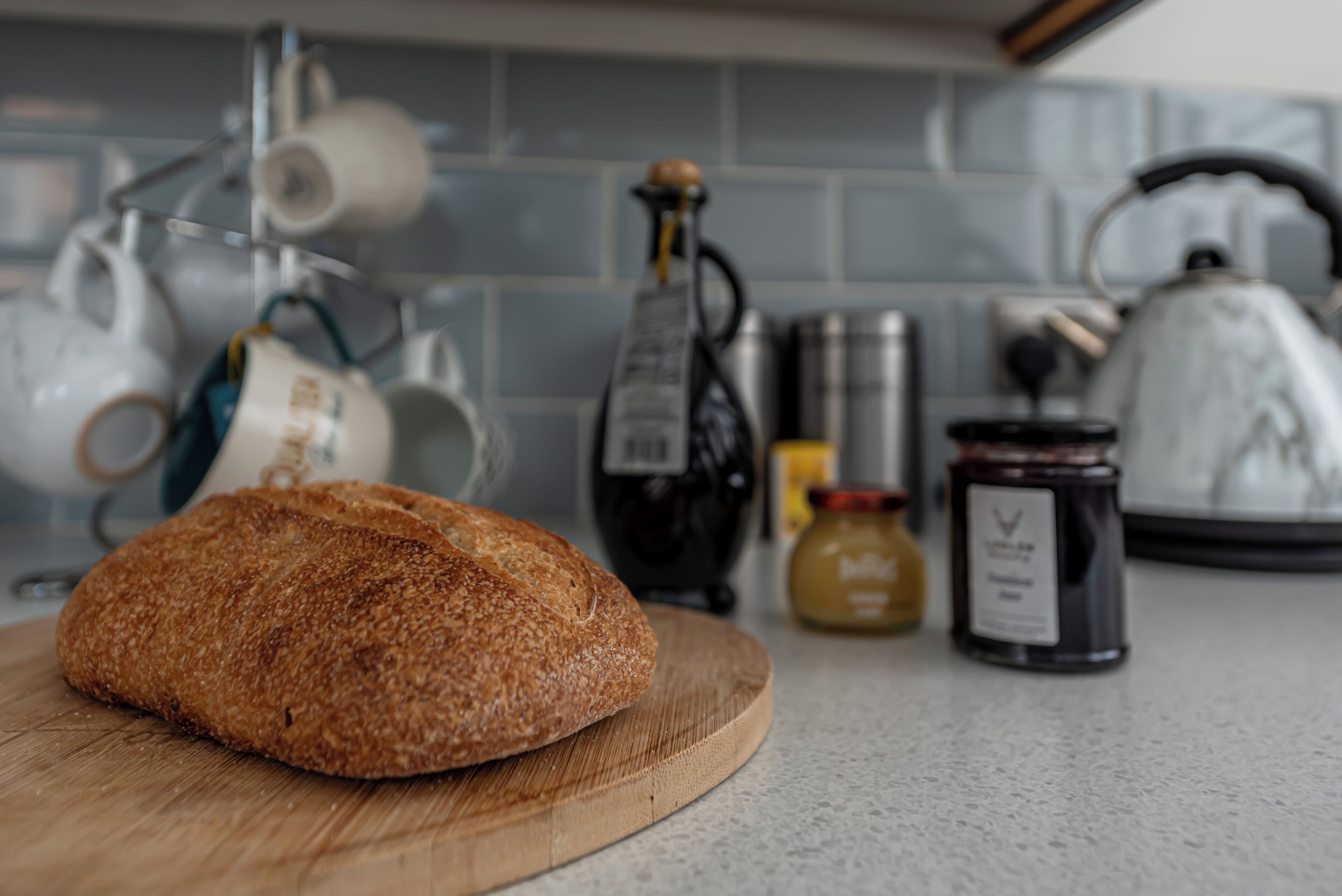 A loaf of bread on a wooden cutting board in a kitchen with tiled backsplash and various kitchen items.