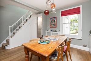 Een eetruimte met een houten tafel, stoelen, een trap, een koelkast, een raam met uitzicht en een schilderij aan de muur.
