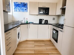 A compact kitchen with a washing machine, a microwave, a coffee maker, and a stove. - River Views | 2BR 2BA | Roof Terrace + Parking - Pass the Keys (London)