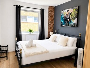 A modern bedroom with a black metal bed frame, white bedding, a window with curtains, a brick wall, a painting, and a radiator. - River Views | 2BR 2BA | Roof Terrace + Parking - Pass the Keys (London)