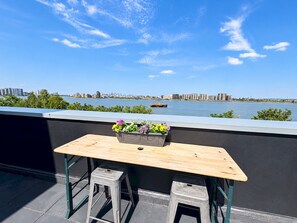 A balcony with a wooden table and two stools, overlooking a river with buildings in the distance. - River Views | 2BR 2BA | Roof Terrace + Parking - Pass the Keys (London)