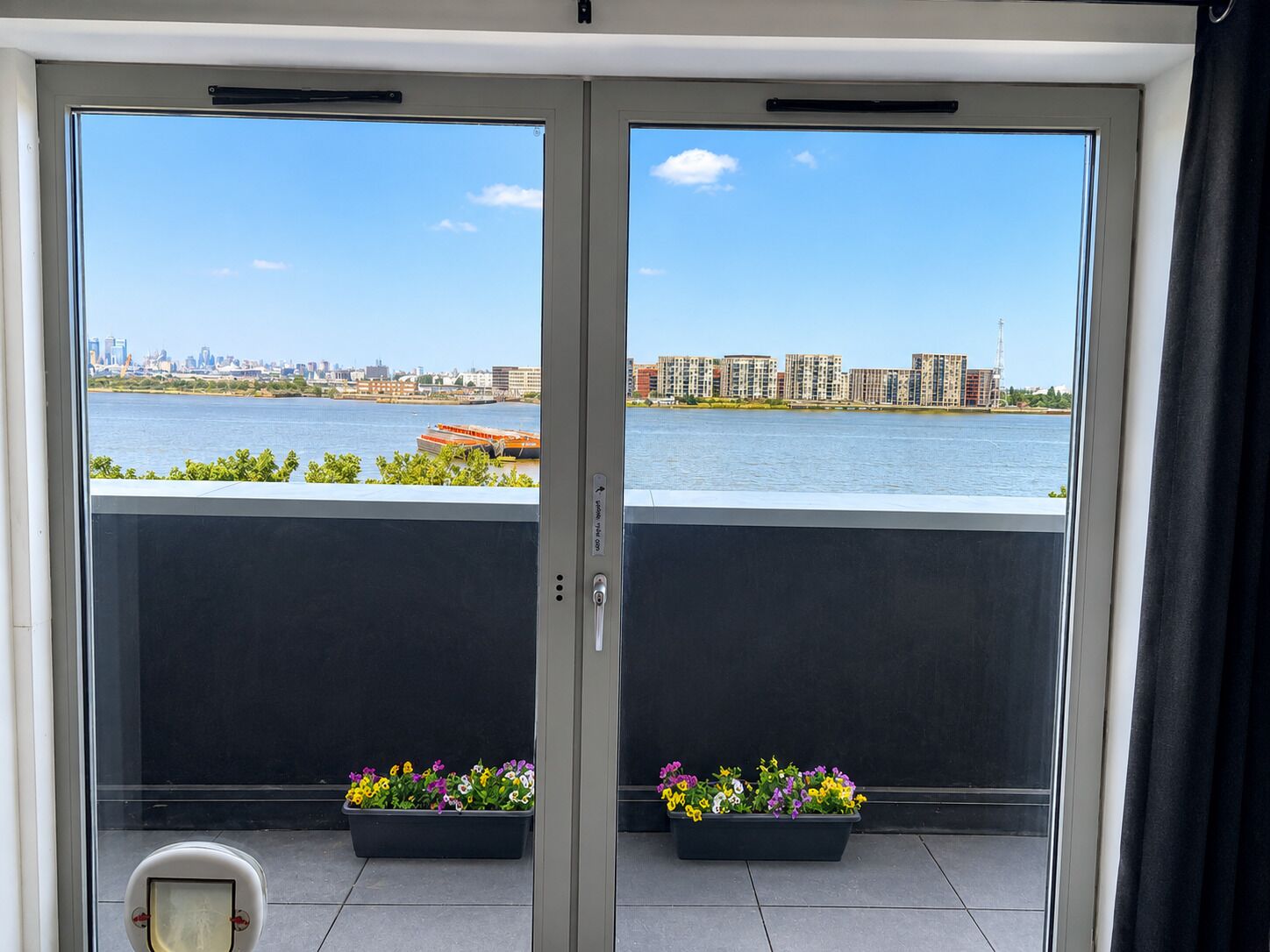 A room with a view of a river and buildings across it.