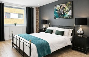 Un dormitorio con una cama, dos mesitas de noche, una ventana con cortinas, un radiador y un cuadro colgado en la pared.