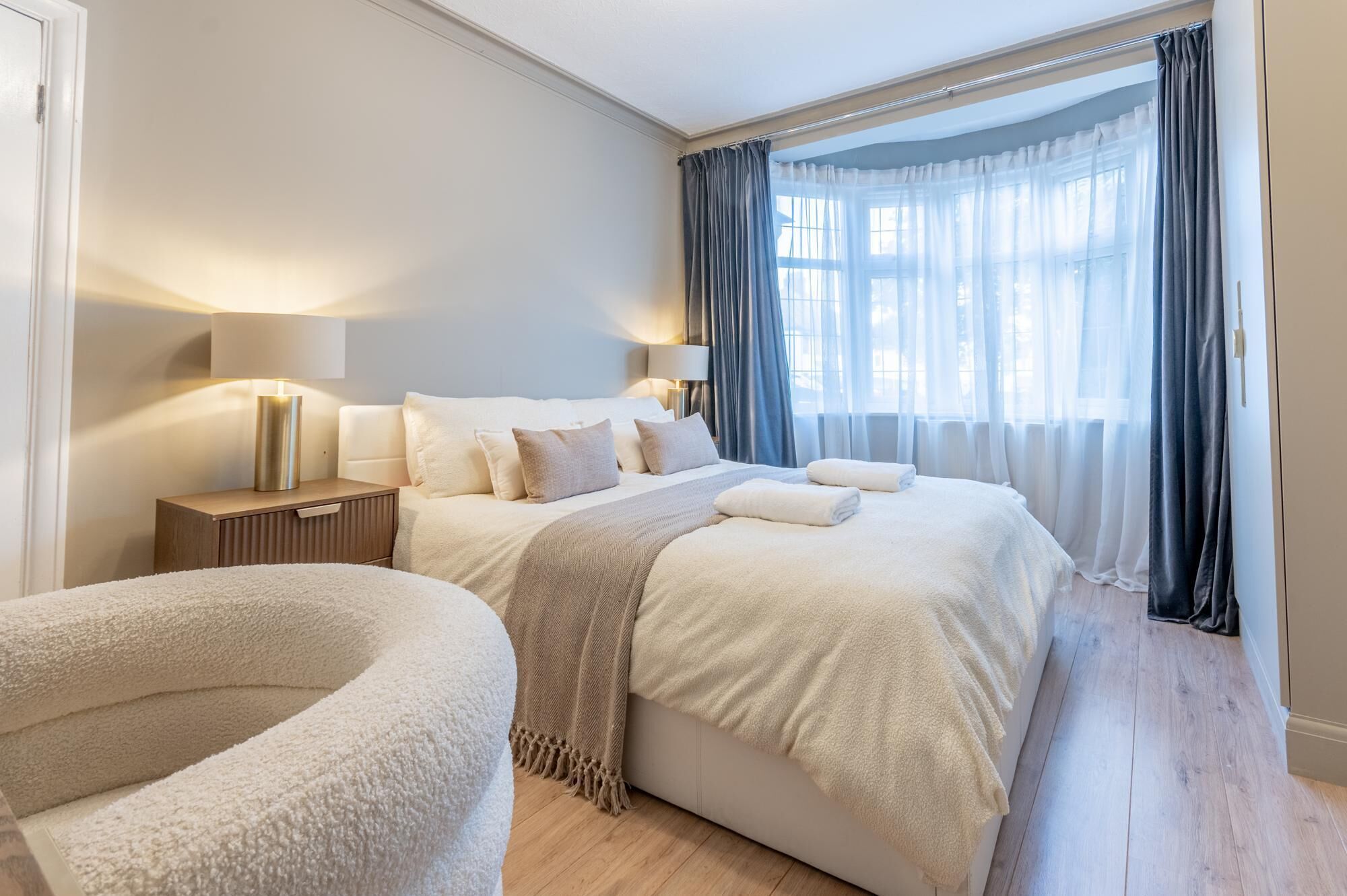 Habitació d'hotel amb un llit gran, tauletes de nit, làmpades, una cadira mullida i una finestra amb cortines.