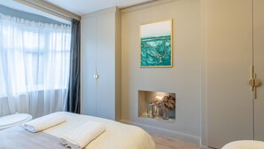 A hotel room with a bed, a painting, a small shelf with decorative items, and a window with curtains. - Luxury 4-Bedroom house near Southend - Pass the Keys (Southend-on-Sea)