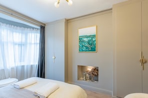A hotel room with a bed, a painting, a small shelf with decorative items, and a window with curtains. - Luxury 4-Bedroom house near Southend - Pass the Keys (Southend-on-Sea)