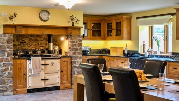A kitchen with a stone fireplace, wooden cabinets, and a dining area with a table set for two. - Inviting Holiday Cottage in Beaworthy (Beaworthy)