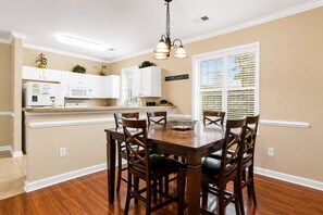 A dining area with a wooden table and chairs, white cabinets, and a window with blinds. - Willow Bend At Barefoot 1111 (North Myrtle Beach)