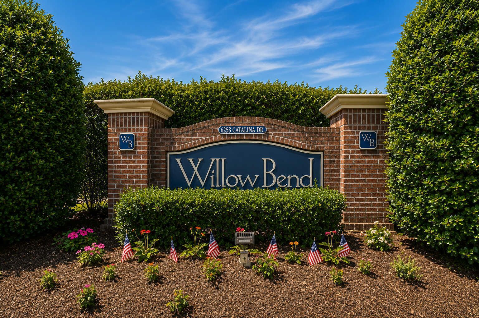 A sign for Willow Bend with a brick archway, surrounded by trimmed hedges and American flags.
