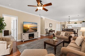 A living room with a fireplace, a ceiling fan, and a television displaying a beach scene. - Willow Bend At Barefoot 1111 (North Myrtle Beach)