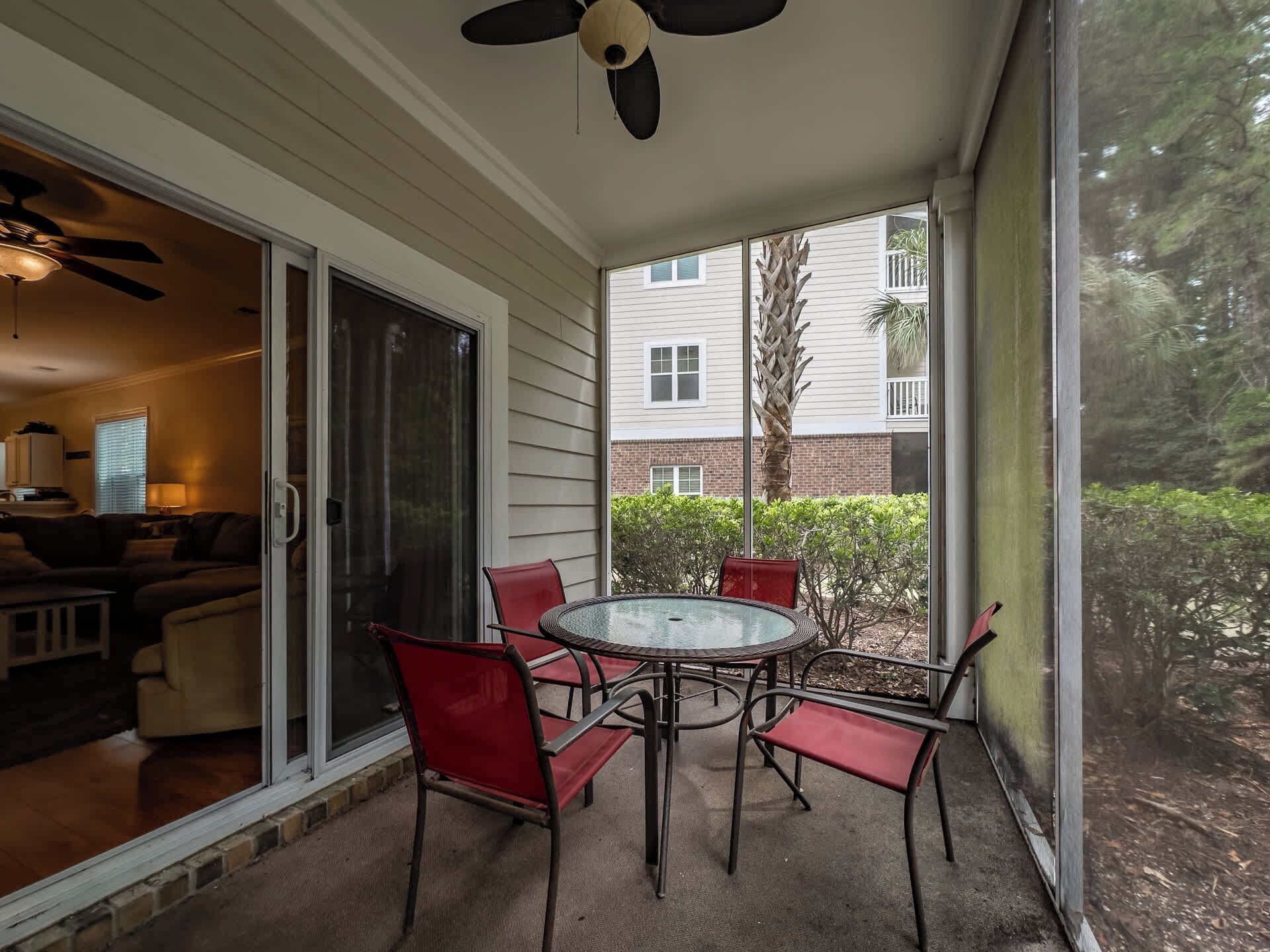 A covered patio with a glass door leading to a living area, a round table with chairs, and a view of a residential building.
