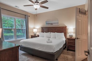 Un dormitorio con cama, mesitas de noche, ventilador de techo, ventana con cortinas y puerta con perchero.
