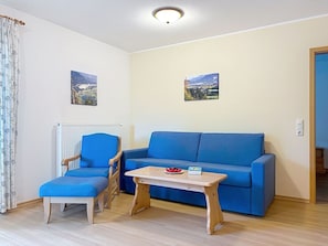Ein Zimmer mit einem blauen Sofa, einem passenden Sessel, einem kleinen Holztisch mit einem Buch und Beeren sowie zwei Landschaftsbildern an der Wand.