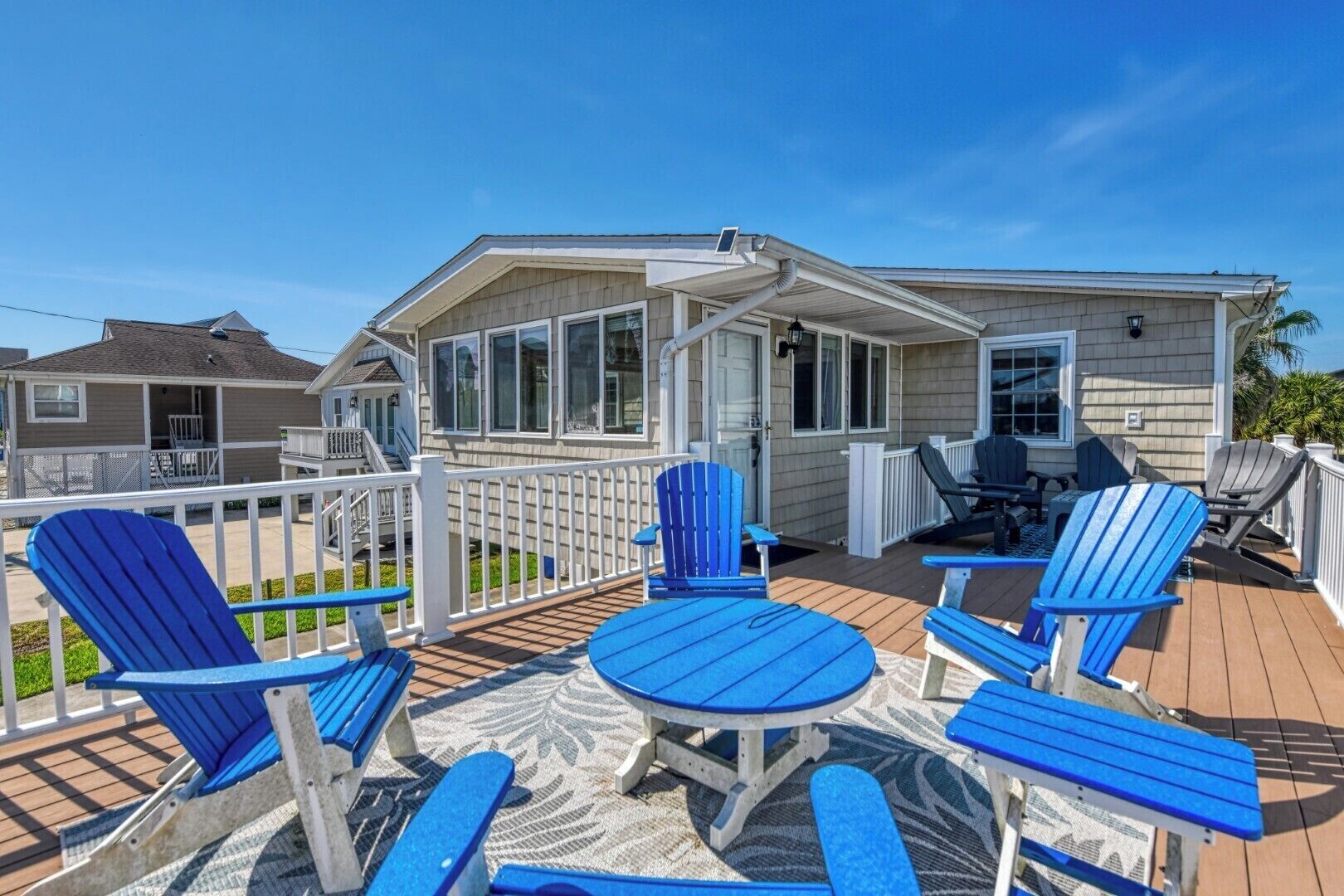 A covered deck with Adirondack chairs and a table, a house with a covered porch, and a clear blue sky.
