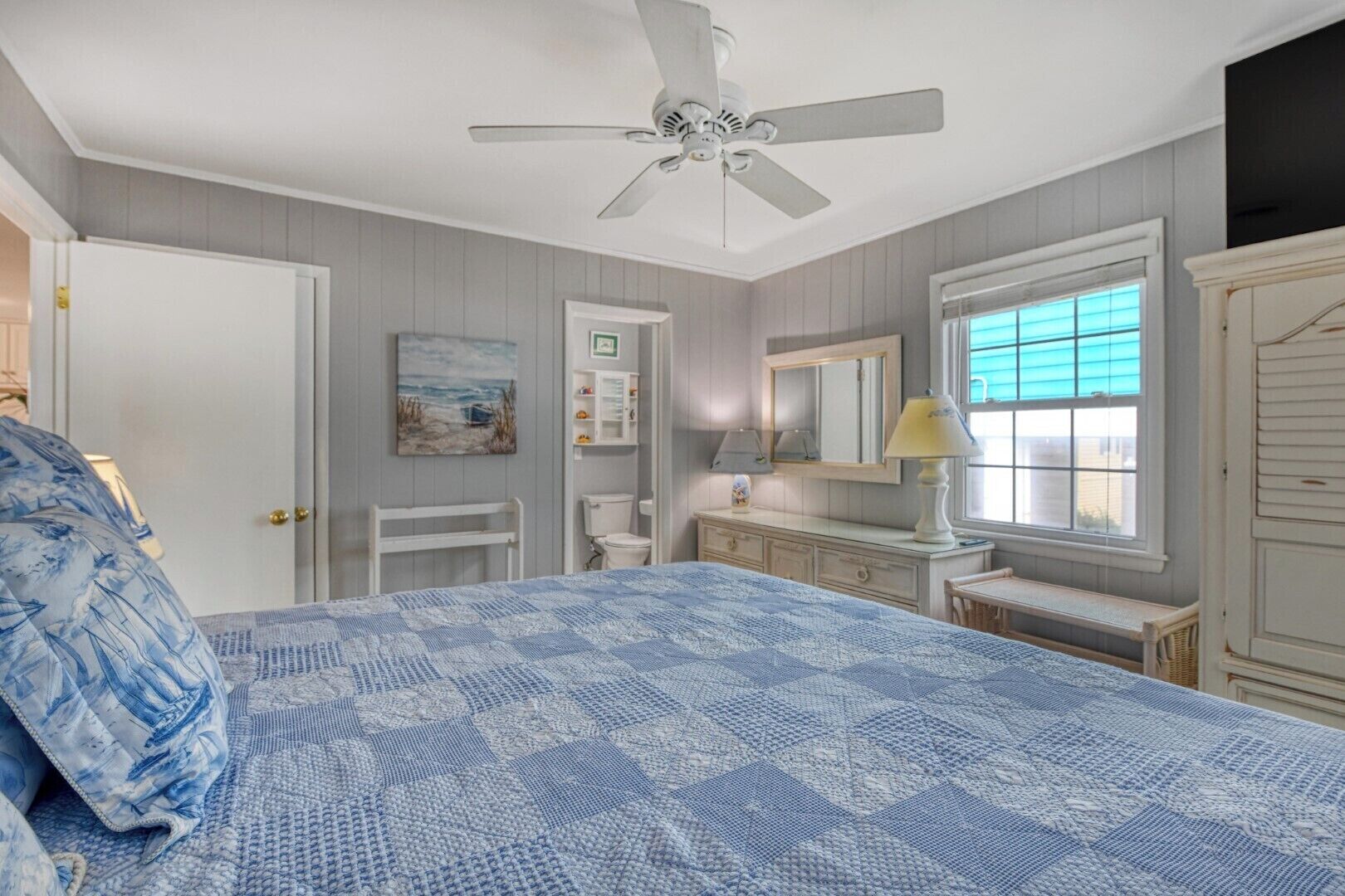 A bedroom with a bed, a ceiling fan, a window with blinds, a bedside table with a lamp, and a dresser.