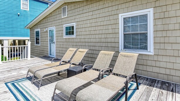 A covered outdoor area with four lounge chairs and a small table, a wooden deck, and a building with beige siding and a blue exterior. - Sail Away - 4 Bedroom, 2.5 Bath Beach Home - Dog Friendly with Fenced In Yard (Garden City Beach)