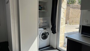 Une buanderie équipée d’un lave-linge, de plusieurs étagères où sont rangés divers objets, et offrant une vue sur l’extérieur par une fenêtre.