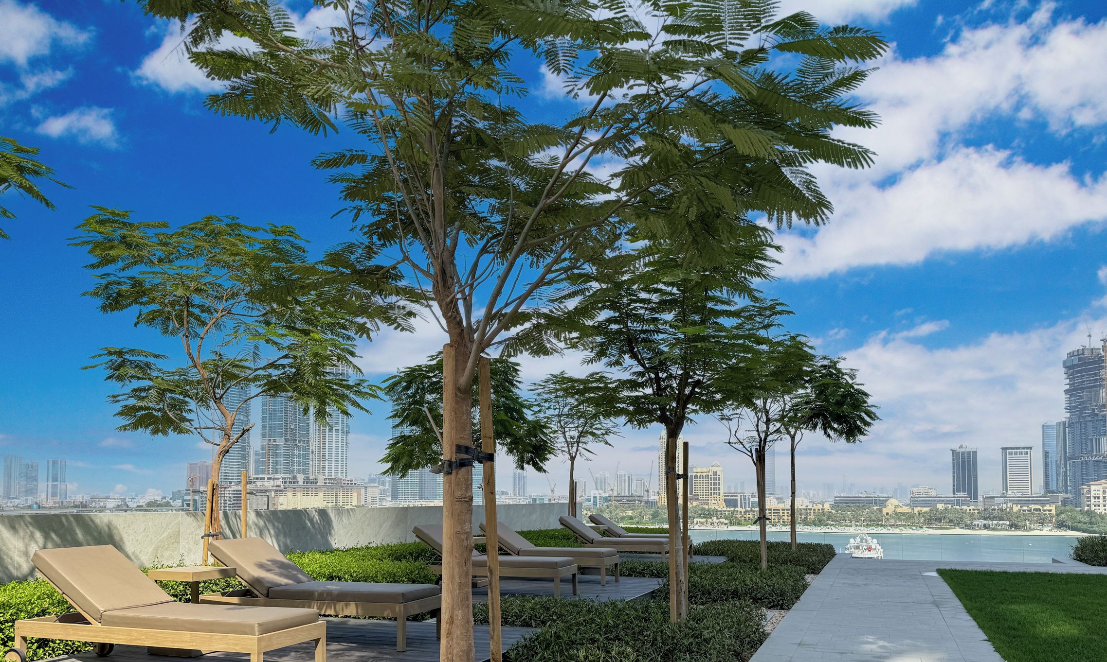 Un espace de détente extérieur moderne, doté de chaises longues et d’un sentier menant à un plan d’eau, avec une vue sur les gratte-ciel de la ville en arrière-plan.