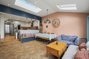 Ein modernes Wohnzimmer mit Fischgrätparkett, einem Holztisch, einem gemusterten Sofa und einem Essbereich mit Tisch und Stühlen.