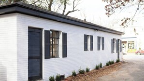 Ein weißes Gebäude mit schwarzen Fensterläden, einem Mauerwerkfundament und einem kleinen Gartenbereich mit Sträuchern.