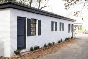 A white building with black shutters, a brick foundation, and a small garden area with shrubs.