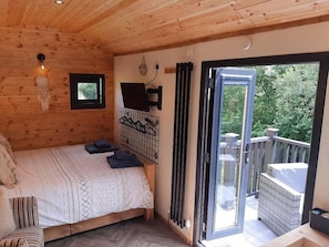 A wooden cabin room with a bed, a small window, a wall-mounted TV, and a sliding glass door leading to an outdoor balcony.