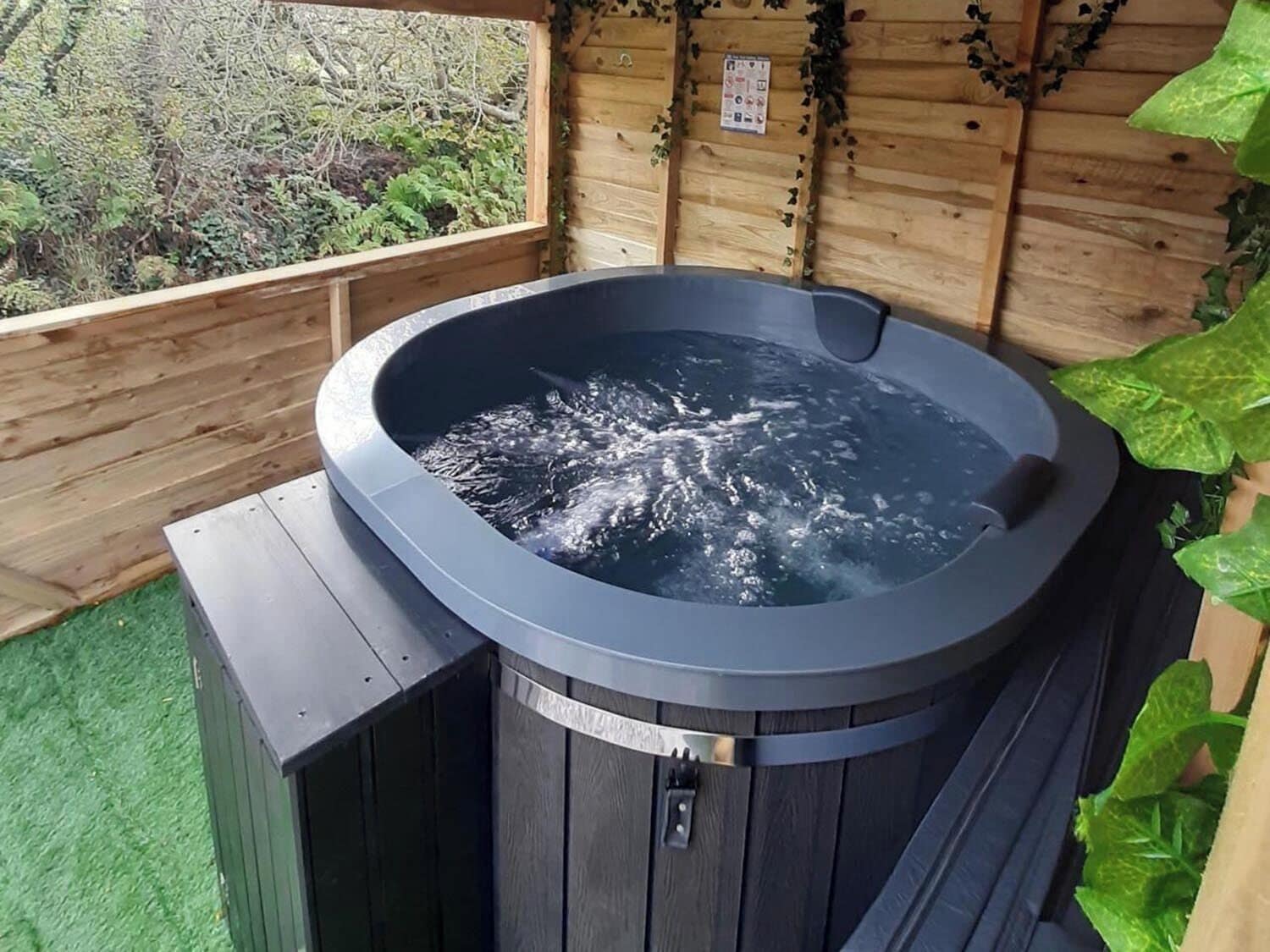 Un jacuzzi con revestimiento de madera, rodeado por una terraza de madera y un muro de madera con follaje.