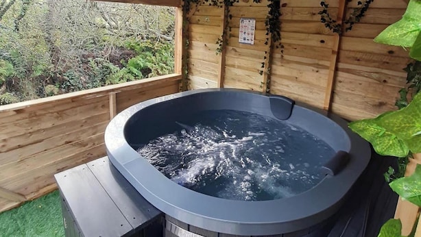 A hot tub with a wooden exterior, surrounded by a wooden deck and a wooden wall with greenery. - Min yr Afon Cabin Sleeps2 Hot Tub Parking (Holyhead)