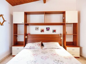 A bedroom with a wooden headboard, a bed with floral bedding, two bedside tables, and a built-in shelving unit with artwork. - Lovely apartment for 3 people with A/C (Imperia)