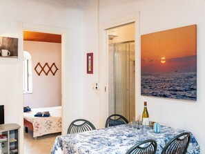A room with a bed, a table with a bottle and glasses, a painting of a sunset, and a shower area. - Lovely apartment for 3 people with A/C (Imperia)