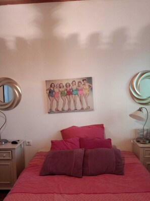A bedroom with a bed, pillows, a nightstand, a lamp, a mirror, and a wall with a poster.