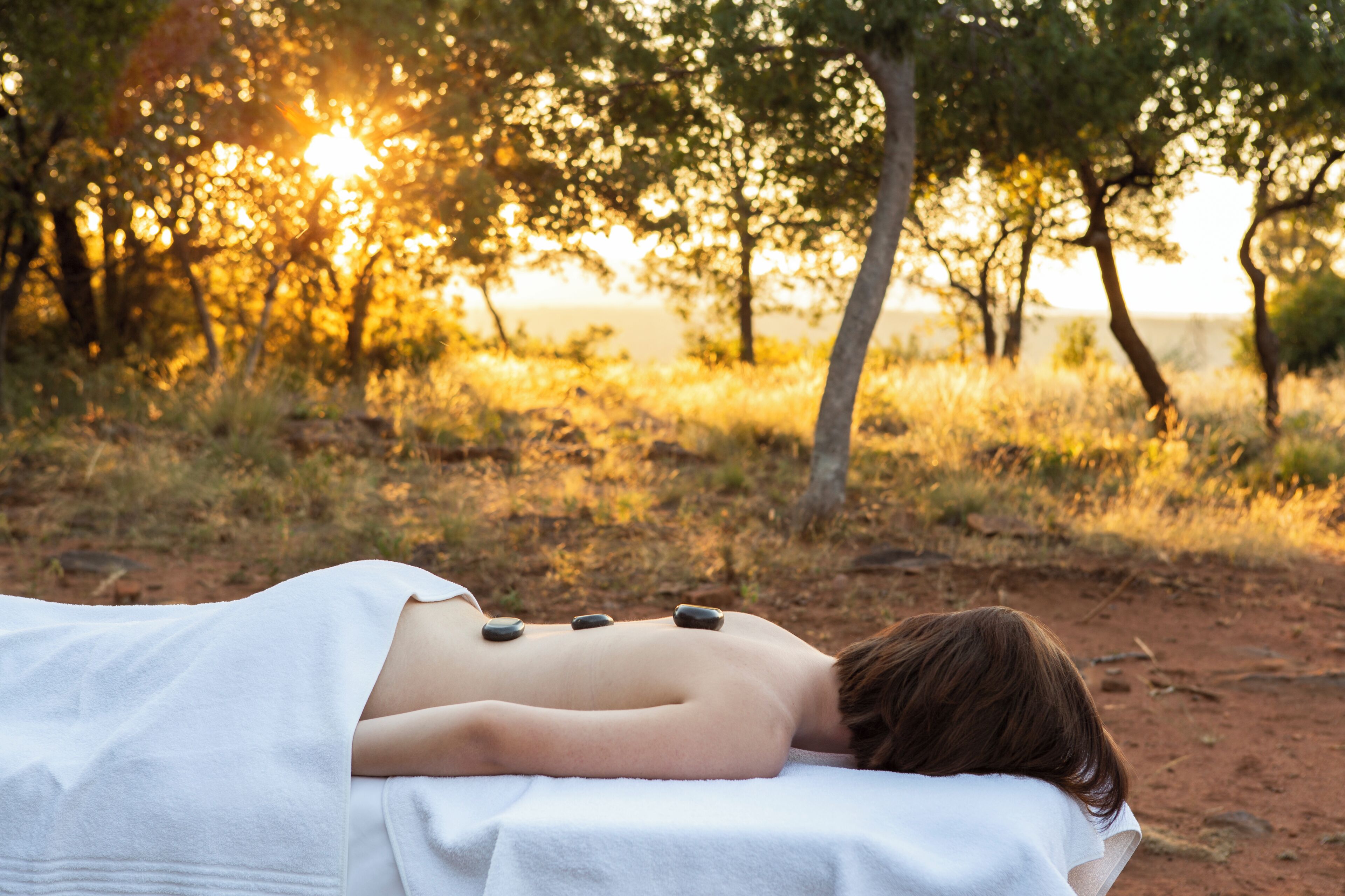 A person receiving a back massage outdoors with stones on their back, surrounded by trees and a sunset in the background.