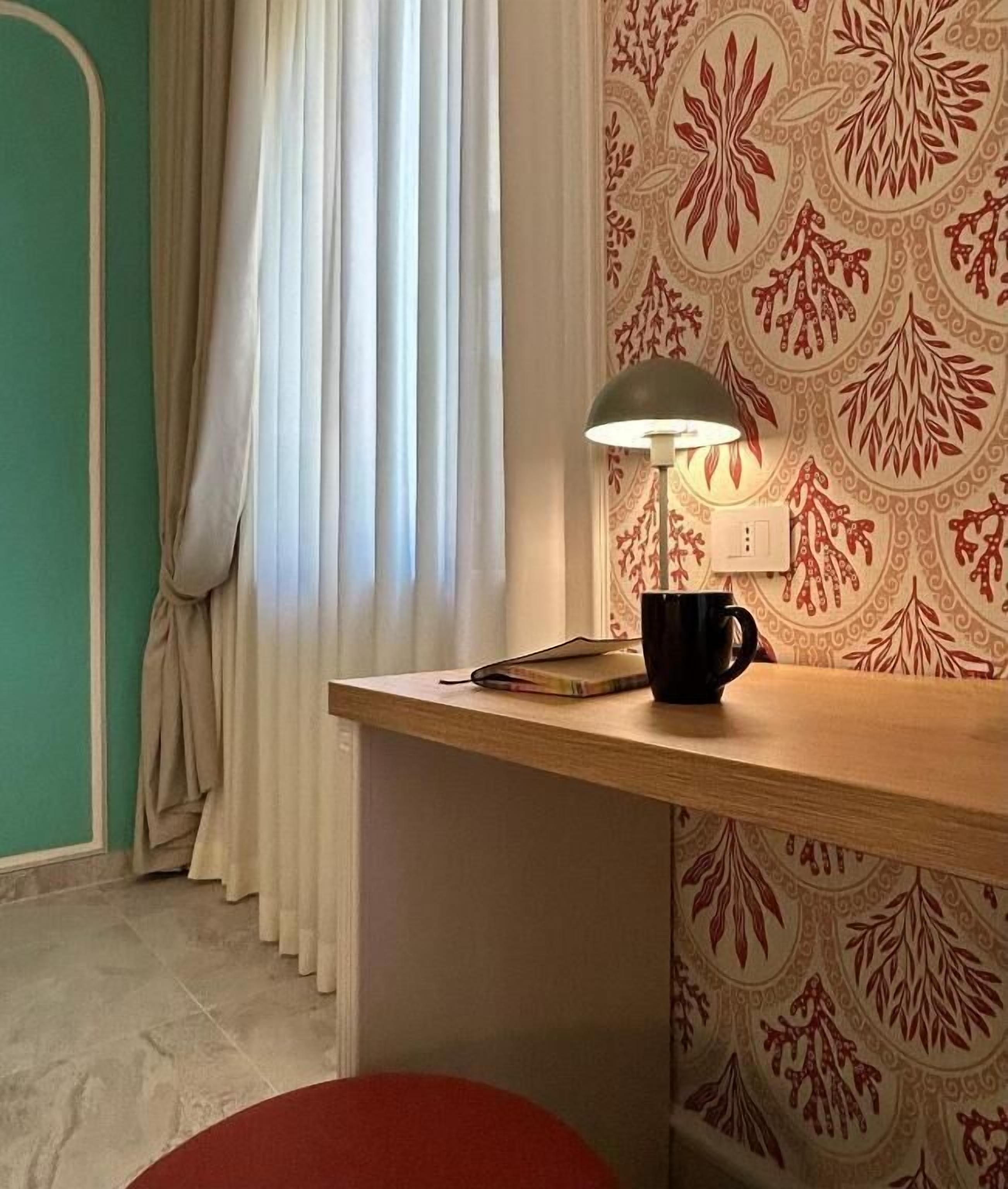 Ein Zimmer mit einem Holztisch, einer Lampe, einer schwarzen Tasse und einem gemusterten Wandteppich.