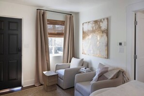 A room with a dark door, a window with beige curtains, a beige sofa, a small wooden stool, and a large abstract painting on the wall.