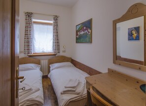 A room with two single beds, a wooden table, a window with patterned curtains, and two paintings on the wall. - Ground floor flat with a small terrace in the heart of San Vito di Cadore (San Vito di Cadore)