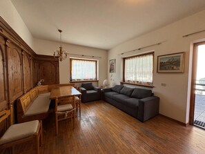 A room with a wooden dining table, a sofa, and a chair. There are windows with curtains, a chandelier, and a painting on the wall. - Ground floor flat with a small terrace in the heart of San Vito di Cadore (San Vito di Cadore)