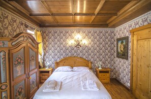 Ein traditionell eingerichteter Raum mit Holzbalkendecke, einem Bett mit weißer Bettwäsche, einem Nachttisch, einer Wandlampe und einer Tür mit einem Gemälde.