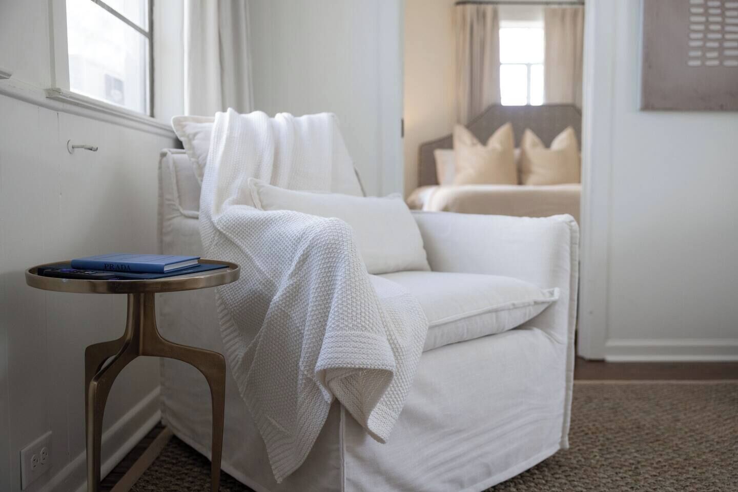 Un sillón blanco con una manta, una mesita redonda con un libro y una vista de una cama al fondo.
