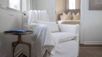 A white armchair with a blanket, a small round table with a book, and a view of a bed in the background.