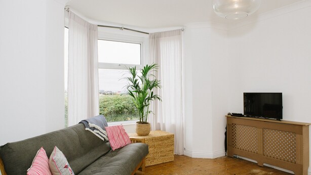 A living room with a sofa, a television on a cabinet, a potted plant, and a view through a window. - Stunning Apartment Near Fistral Beach (Newquay)