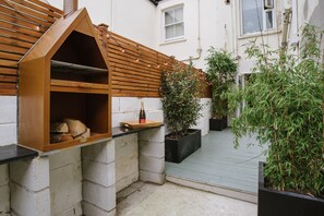 Ein hölzerner Grill für den Außenbereich mit geneigtem Dach, einer Weinflasche und einem Glas auf einer Holzschale.
