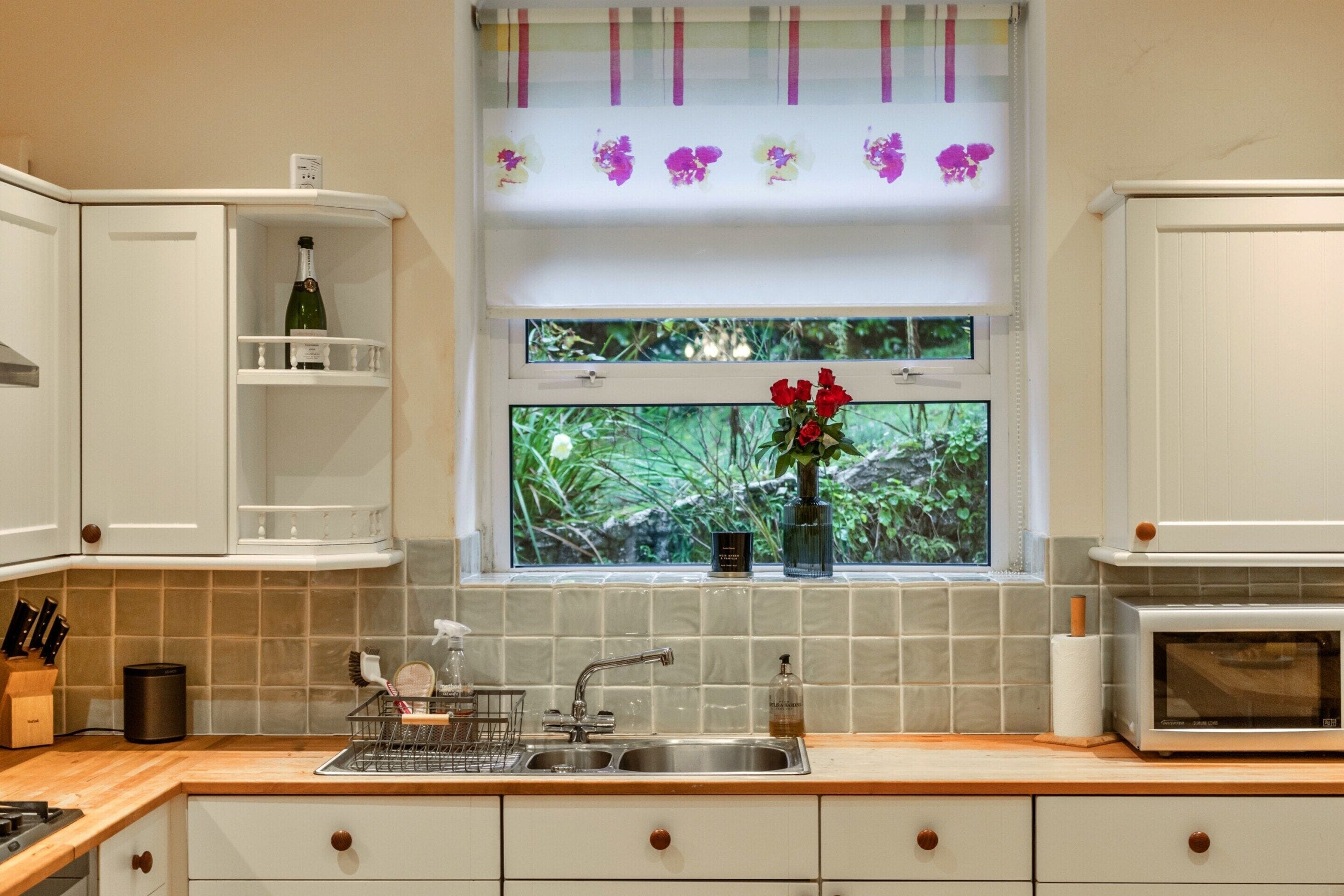 Een keuken met een dubbele spoelbak, witte kastjes, een wijnfles op het bovenste schap en een raam met uitzicht op groen.