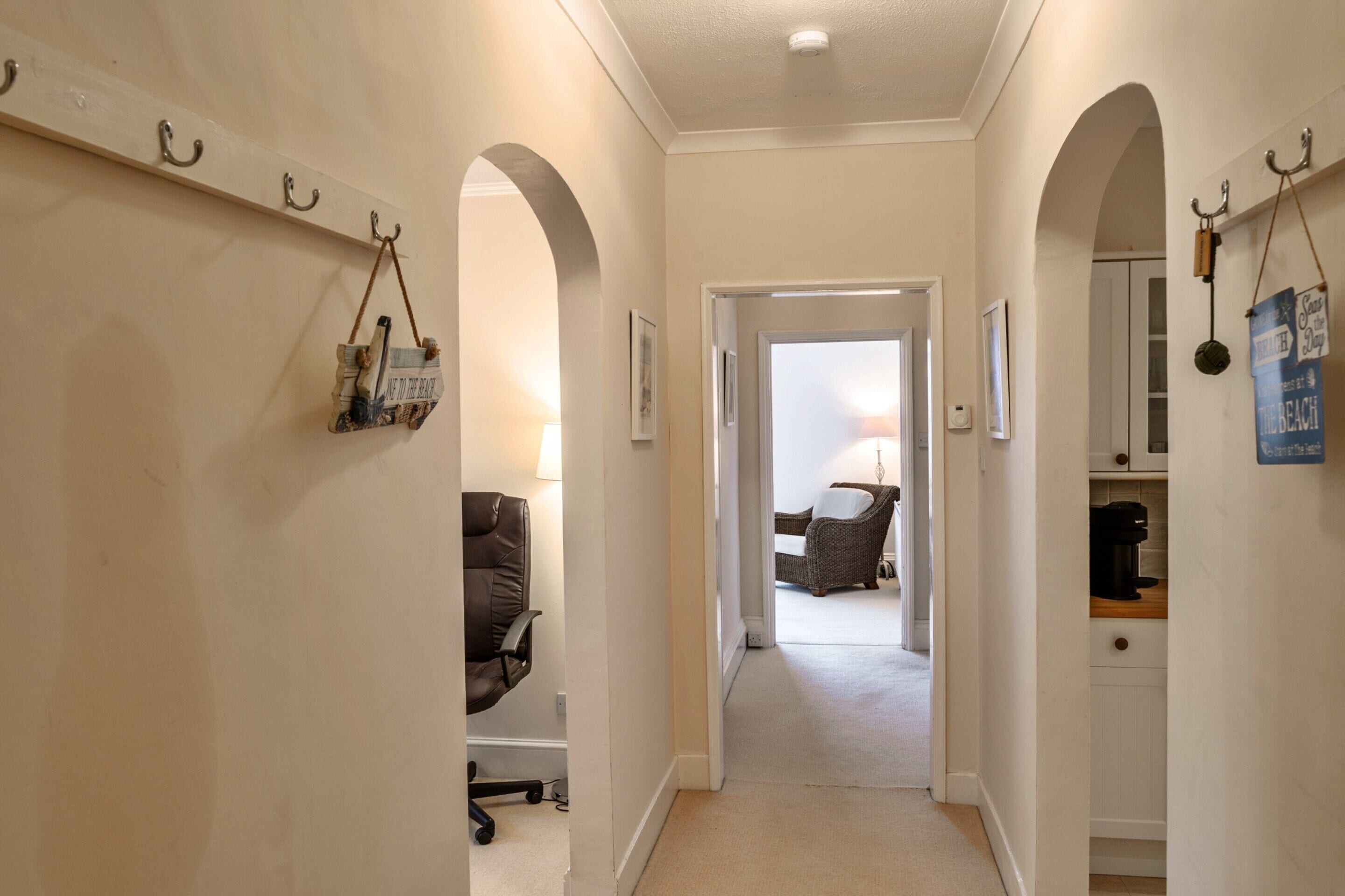 A hallway with a door leading to a room with a chair and a lamp. There are hooks on the wall and a sign with text.