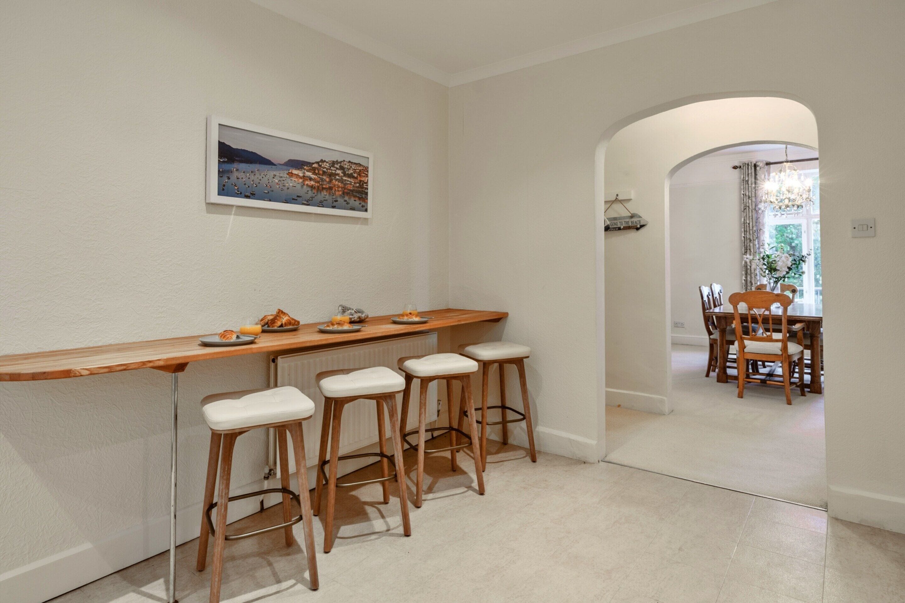 Una cocina con barra de madera, cuatro taburetes, una mesa de comedor con sillas y un cuadro en la pared.
